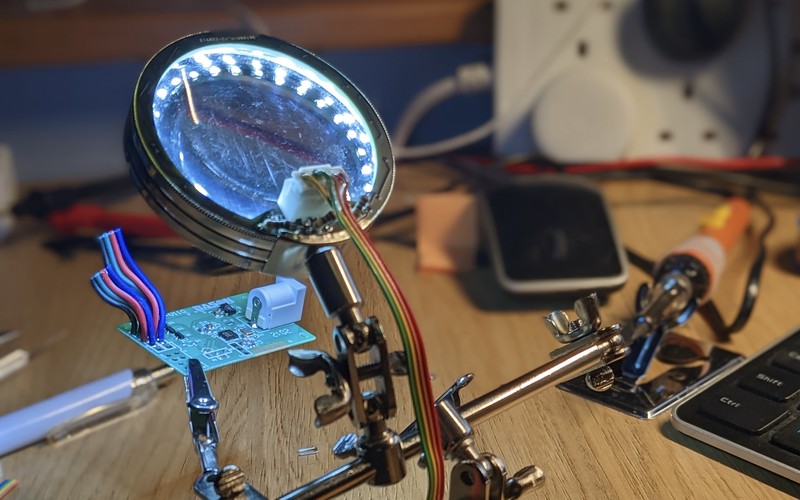 The lit LED ring on its helping hands stand, magnifying glass raised, with the PCB and wiring visible on the workbench